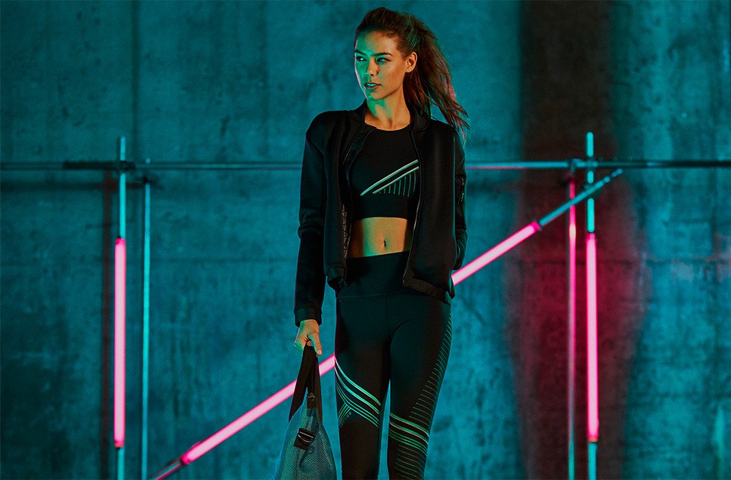 Woman wearing black luminous bra, leggings and bomber jacket carrying grey bag in glowing studio