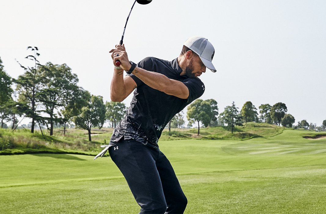 Stephen Curry wearing a UA Range Unlimited golf polo & hat taking a swing on the golf course