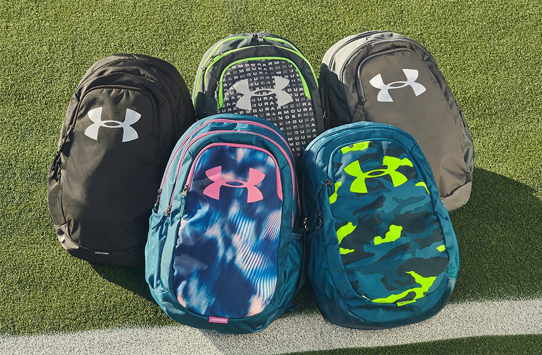 Multiple UA backpacks in a variety of colors & prints laid out together on a turf outdoor field